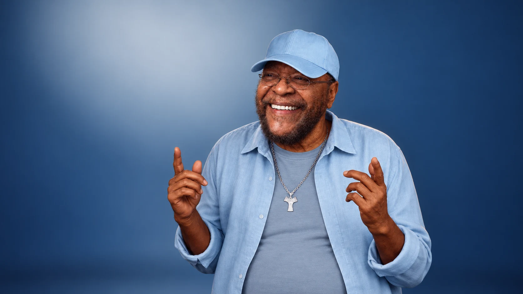 Martinho da Vila sorri, de boné e camisa azul-claro, com as duas mãos erguidas.