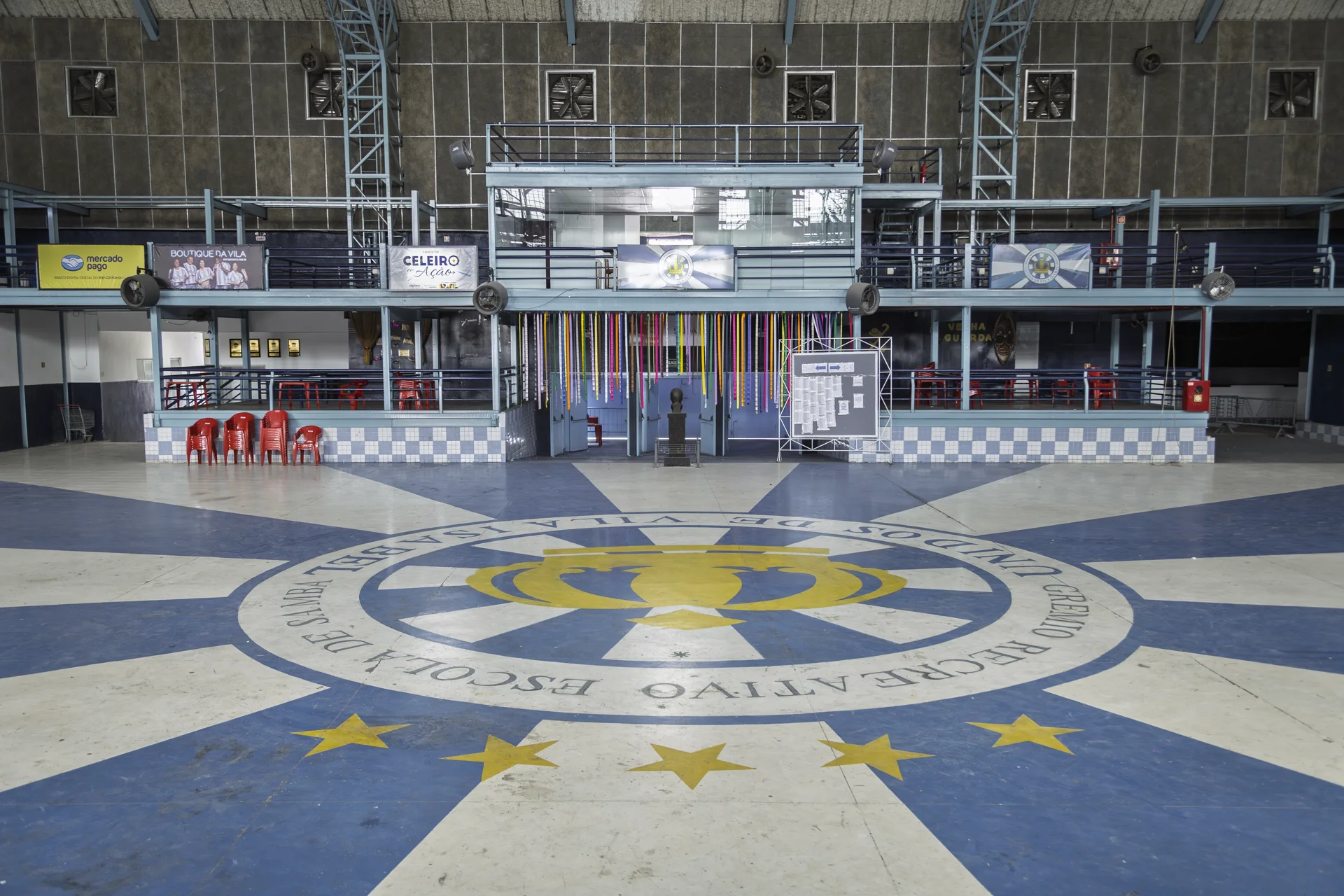 Quadra vazia da Unidos de Vila Isabel: o palco ao fundo, fitas coloridas penduradas e o emblema da escola pintado no chão.